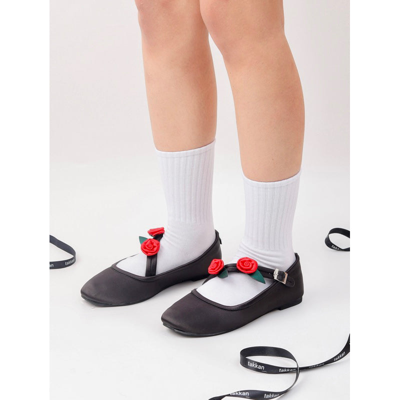 A pair of black flat shoes by Takkan featuring red roses on the strap, worn with white socks.