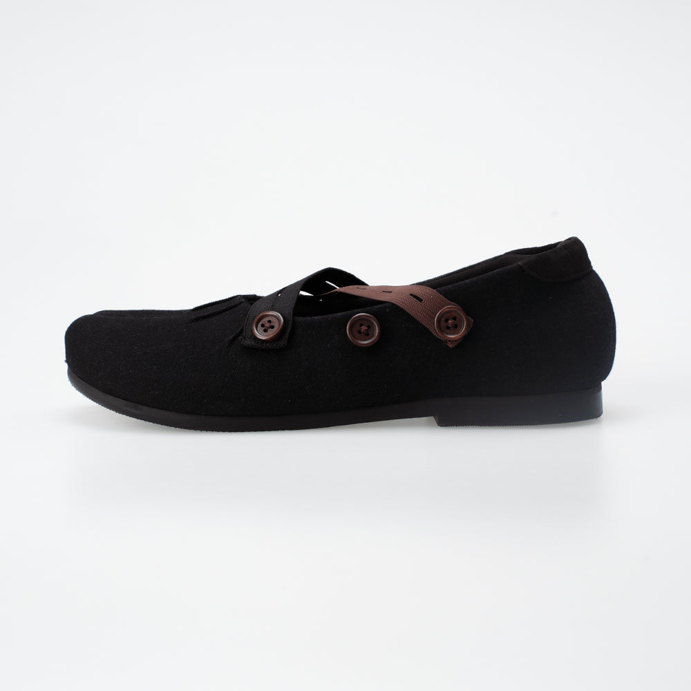 Black flat shoe with brown button accents on a white background