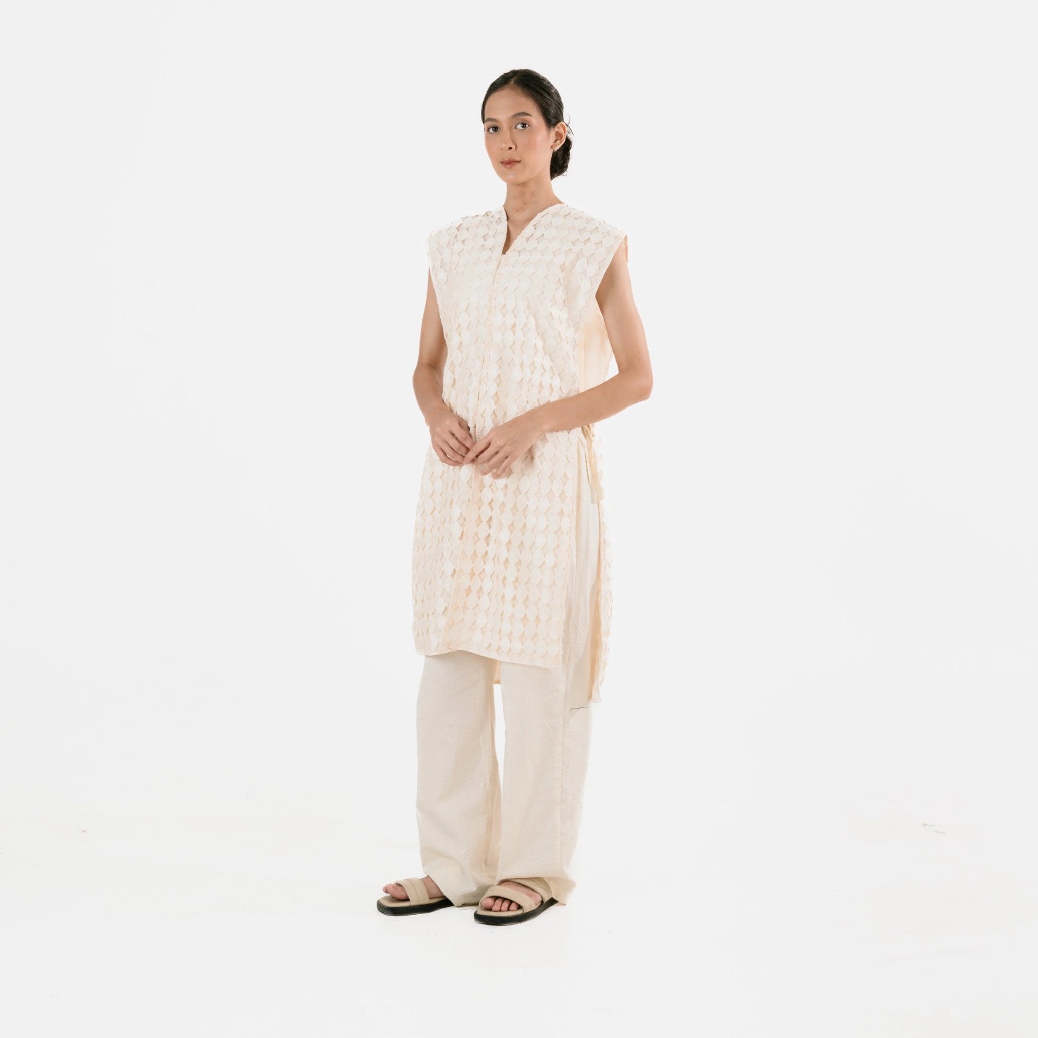 Woman wearing a beige sleeveless long vest by Damakara and pants on a white background