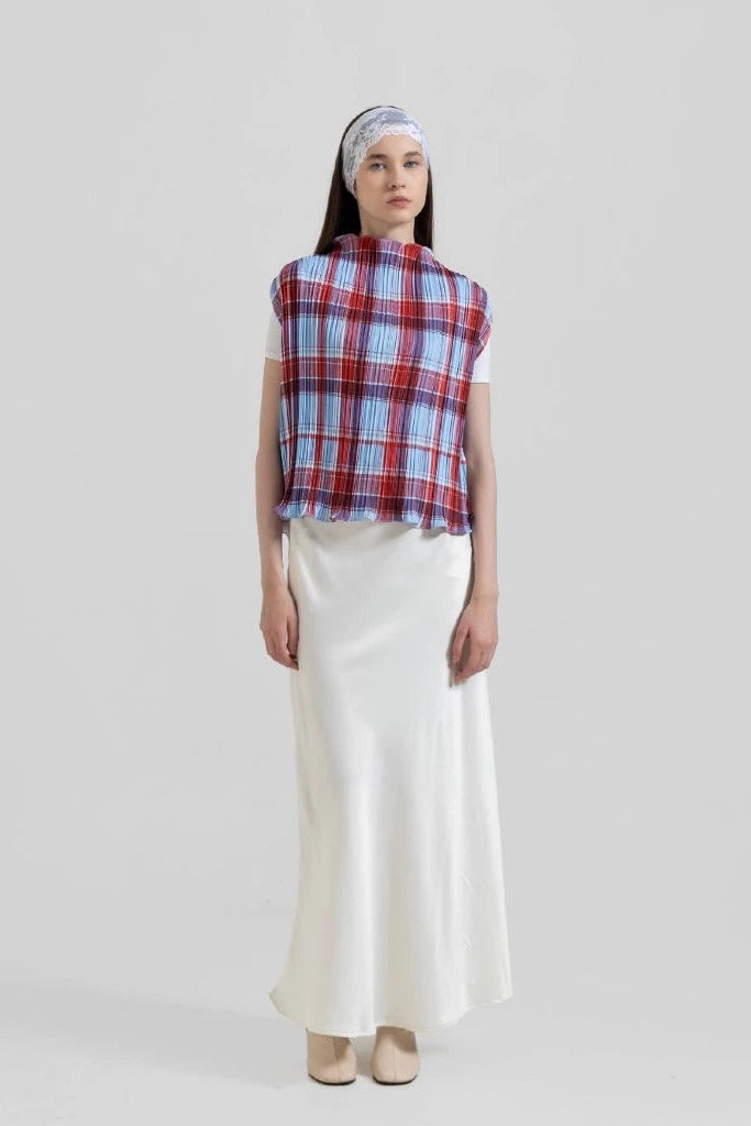 A model stands against a light gray studio background wearing a sleeveless red, blue, and white plaid top by Tuff Puff, a white satin-like maxi skirt, and a patterned headband.