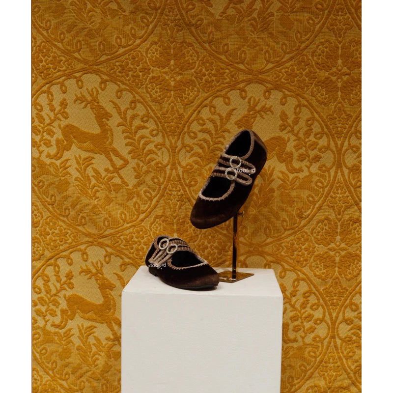 Brown flatshoes with double strap and crystal buckle accents, displayed on a white pedestal against a golden deer-patterned wallpaper background.