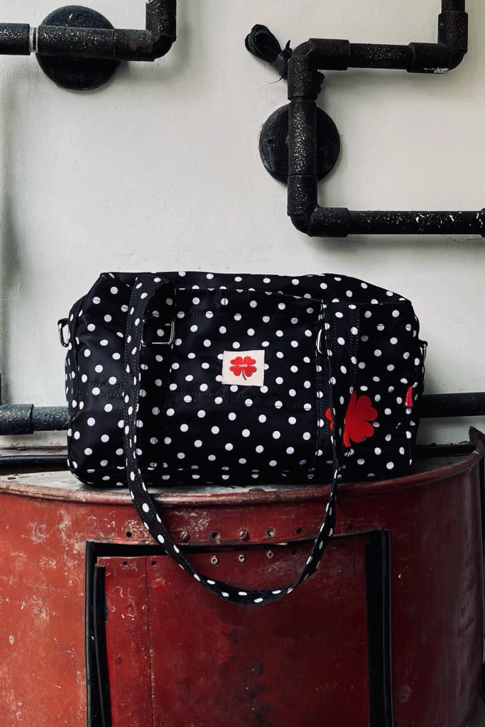 Black polka dot shoulder bag by Luella with red accents on a red metal barrel against a white wall with black pipes.