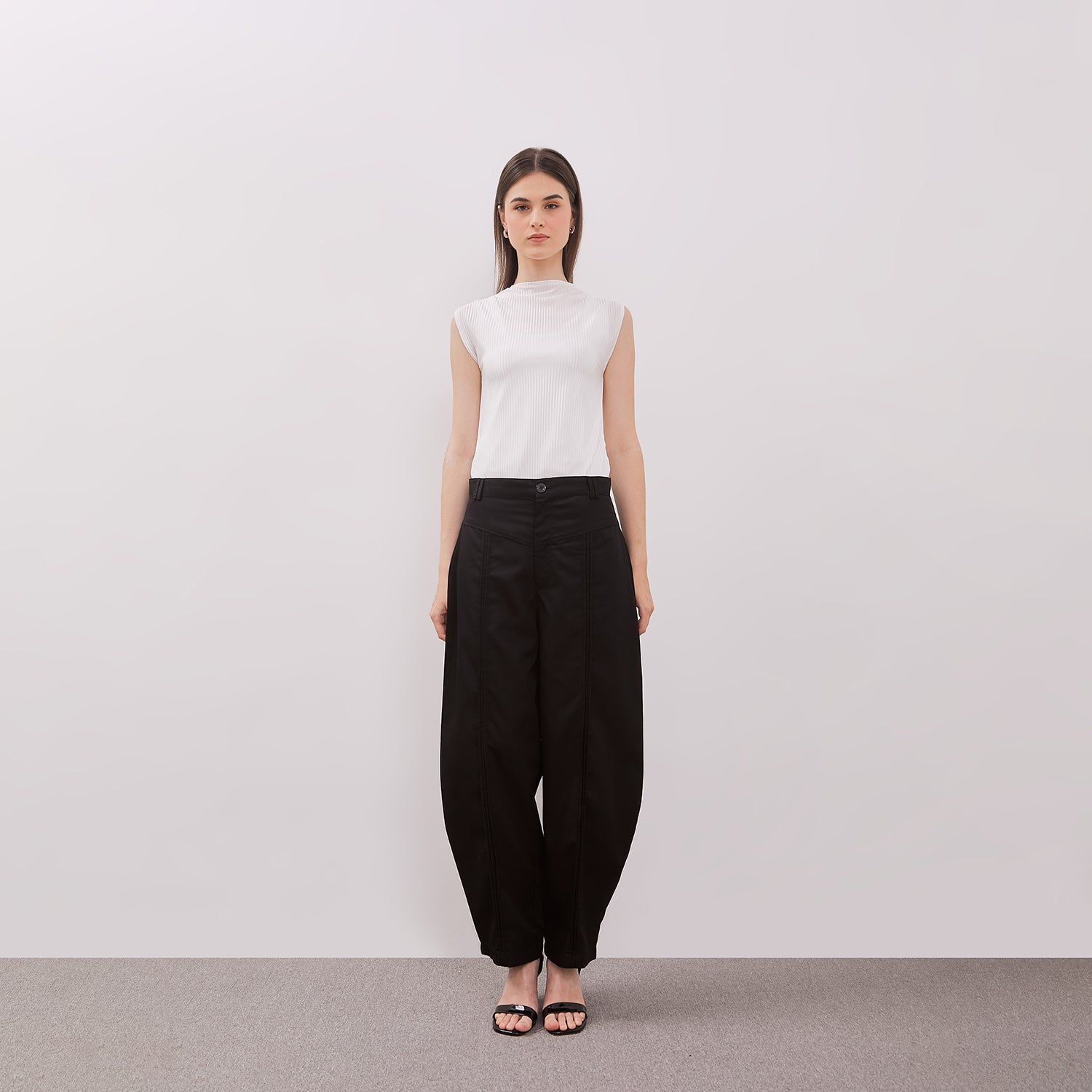 Woman wearing a white sleeveless top and black barrel pants against a plain background