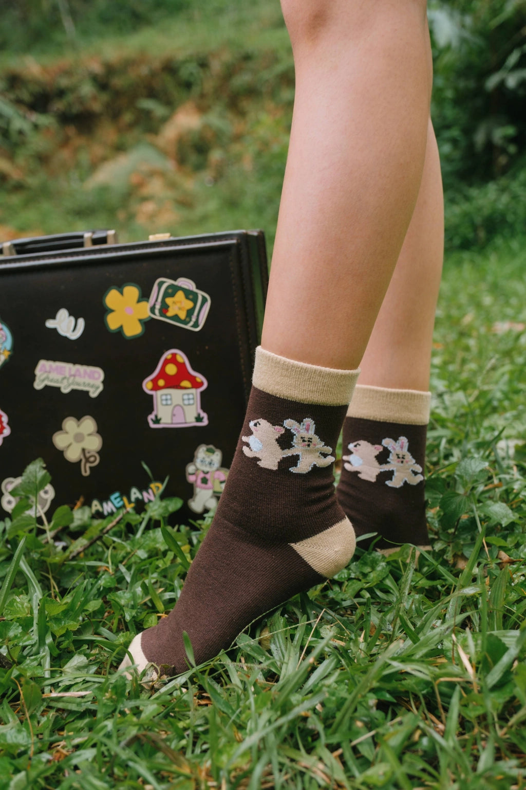 A person's legs standing on green grass next to a black suitcase covered in colorful stickers, wearing dark brown socks by Tome Ame with cream cuffs and white cartoon rabbit patterns.
