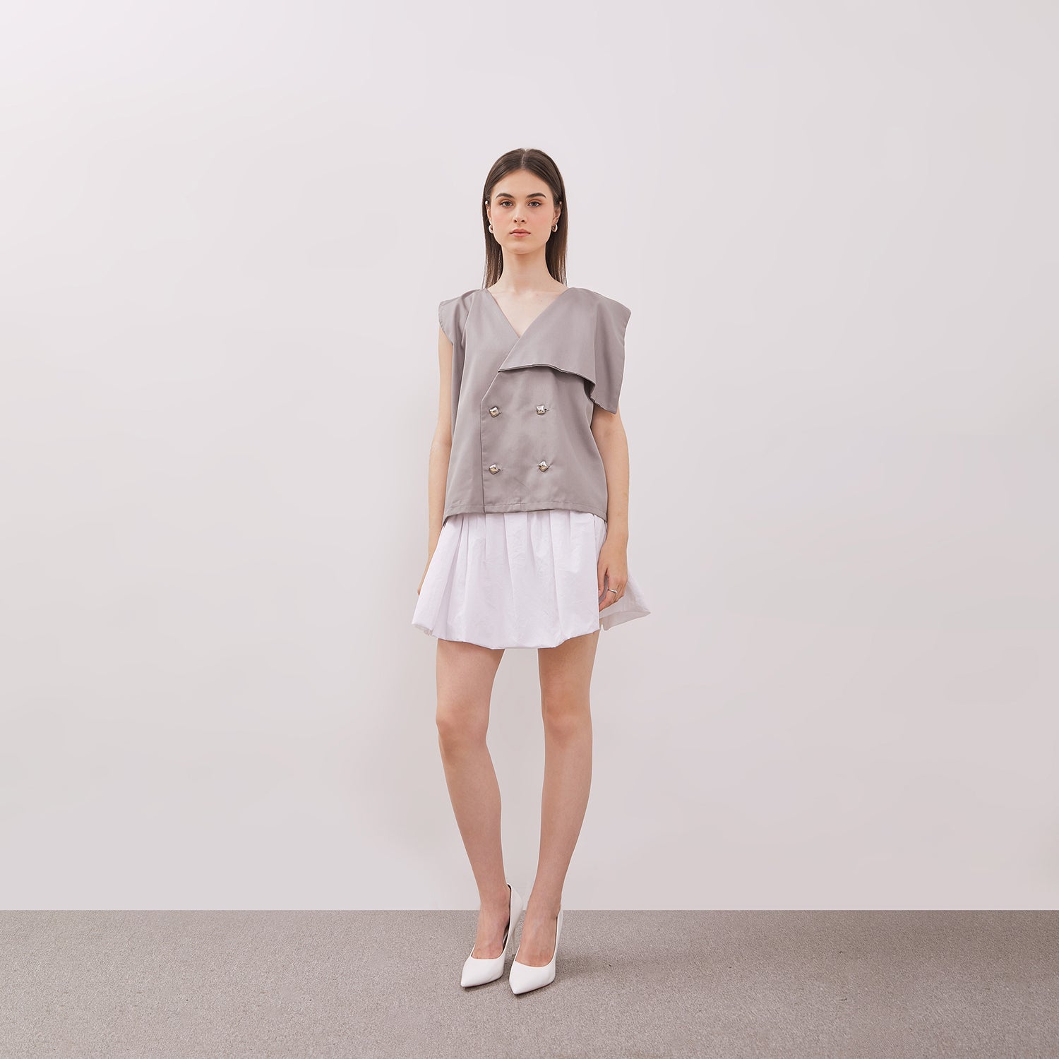 Woman wearing Grey Athena Vest, a gray sleeveless top with buttons, and a white skirt on a plain background