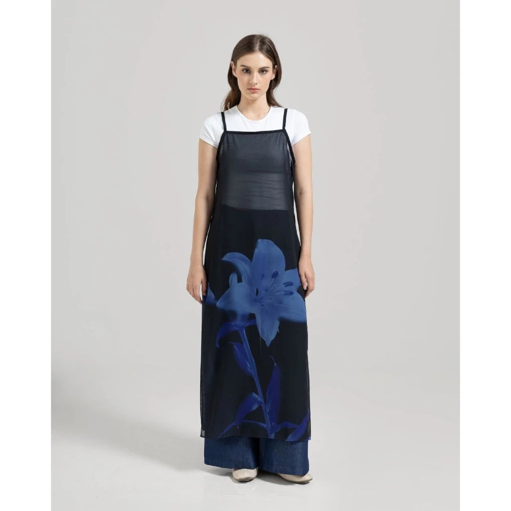 A woman wearing a long black sleeveless maxi dress by Tuff Puff with thin straps and a large blue floral print near the hem, styled over a white long-sleeve turtleneck top. The dress has a loose, flowy silhouette and falls to the floor.