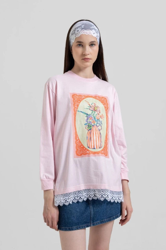 A model wearing a light pink long-sleeve t-shirt by Tuff Puff with a floral graphic print on the front, a white lace hem, and a white lace headband, paired with a denim skirt.