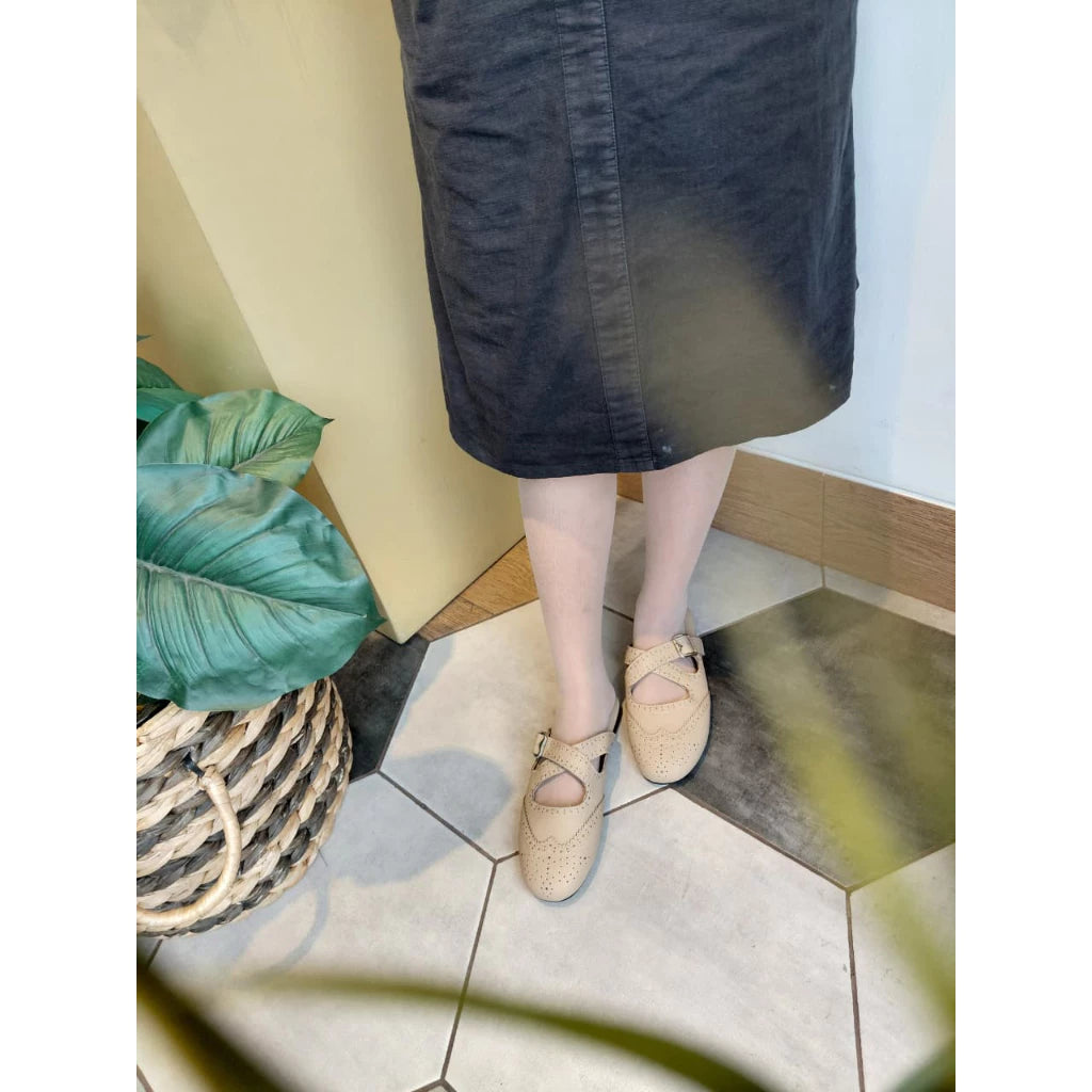 Person wearing a black skirt and cream mules on a tiled floor with a plant in the background