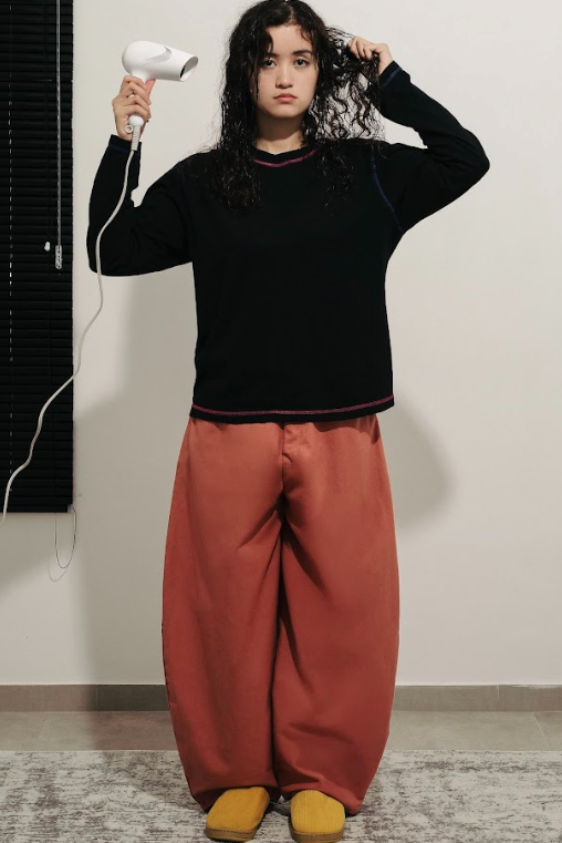Person wearing a black long-sleeve top and dark orange barrel pants standing in a room holding hair dryer.