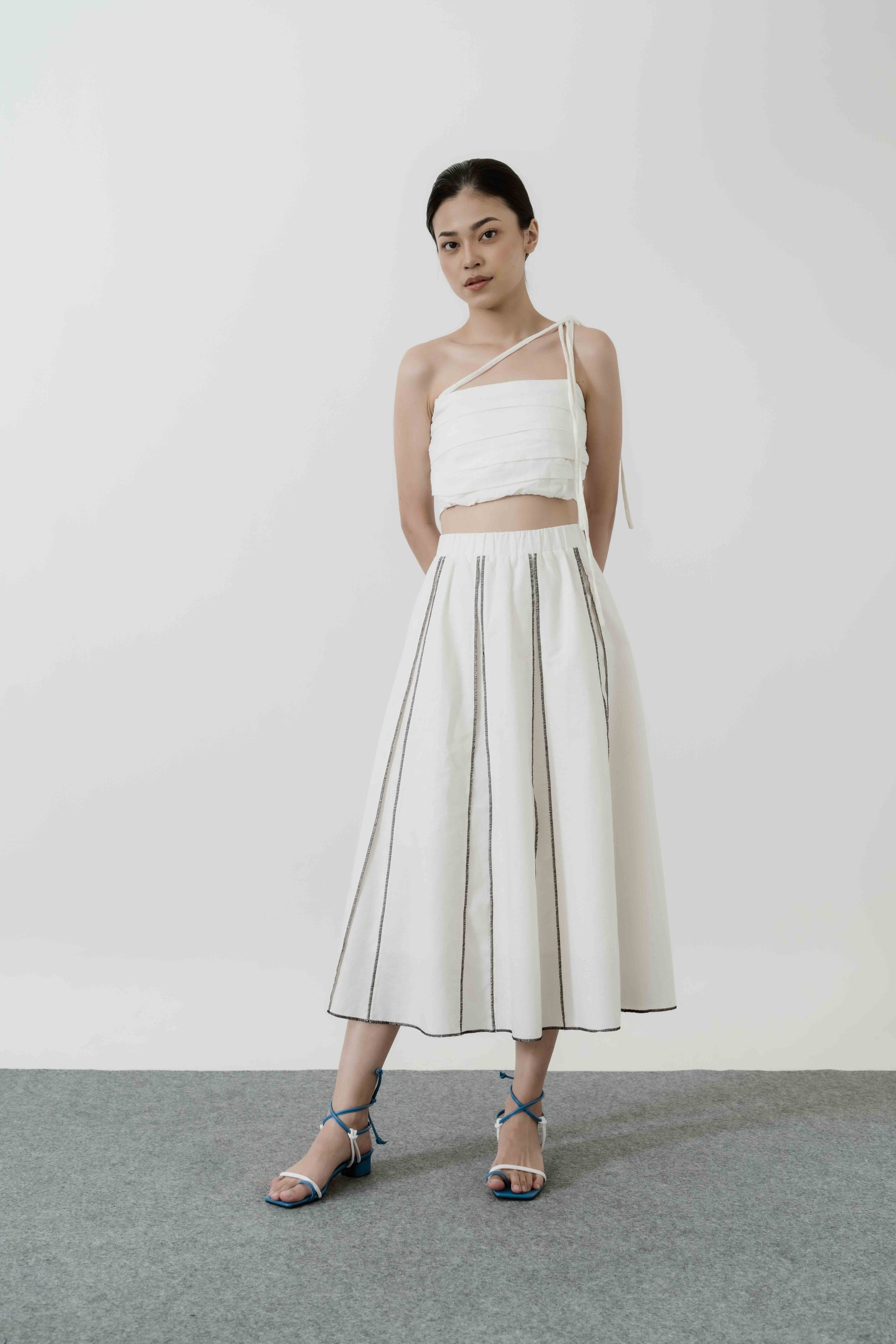 A woman standing wearing a white midi skirt by Morningsol with a flared design, paired with a white top and blue strappy sandals.