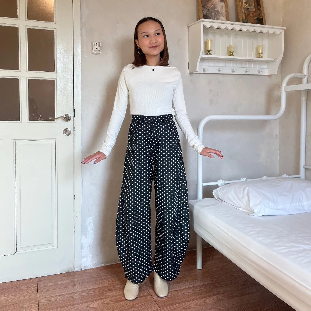 Woman wearing a white top and black polka dot barrel pants standing in a bedroom.
Seorang wanita yang mengenakan atasan putih dan celana barrel bermotif polkadot hitam sedang berdiri di dalam kamar tidur.