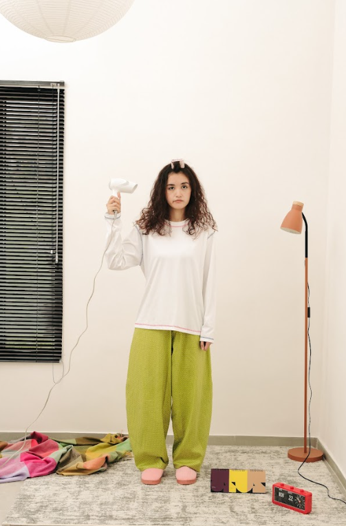 Person wearing a white top and green barrel pants standing in a room holding a hairdryer.