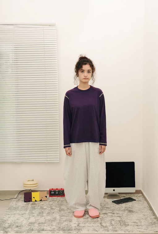 Person wearing a purple sweater and white barrel pants standing in a room with a computer and books on a shelf.