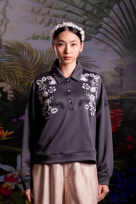 Woman wearing a dark gray embroidered polo shirt with floral patterns by Nona against a tropical background.
