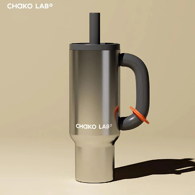 A black Chako Lab mug with a black handle and black straw, placed on a rock with flowers in the background.