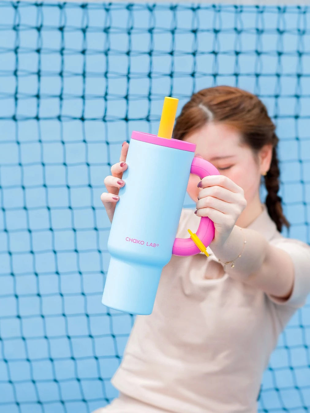 Person holding a blue Chako Lab tumbler with pink lid and yellow straw against a net background.