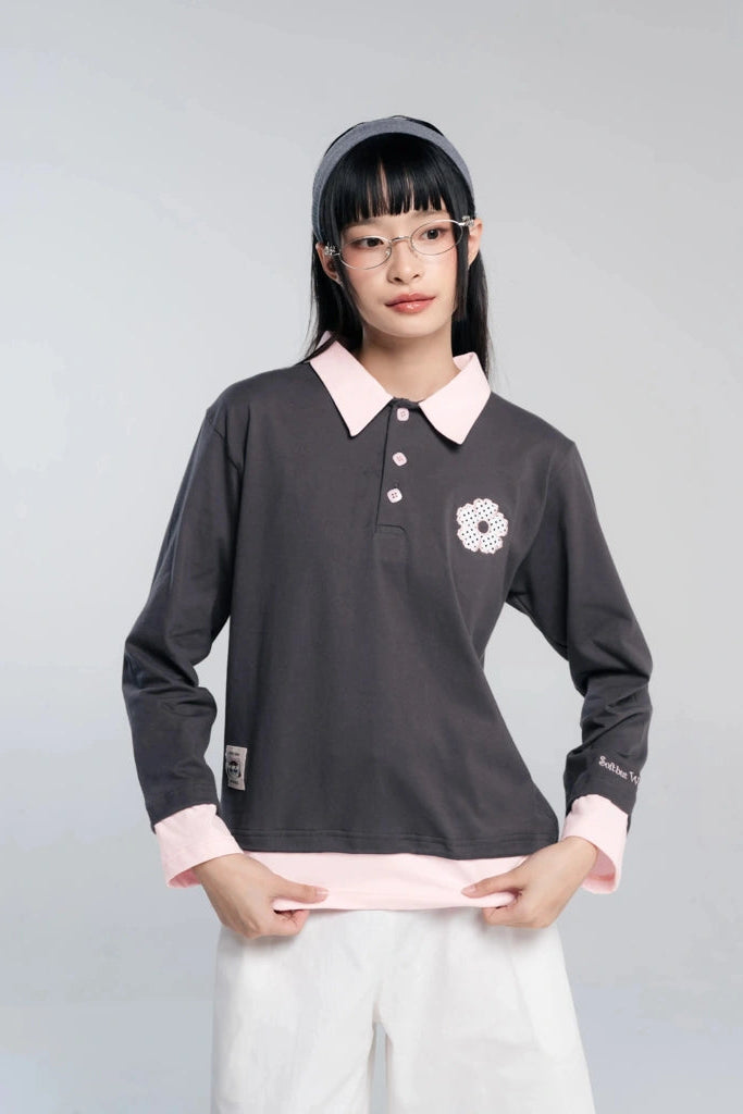 A young woman wearing a dark gray long-sleeve polo shirt by Tome Ame with a light pink collar and cuffs, paired with white pants and a gray headband, against a light gray studio background.