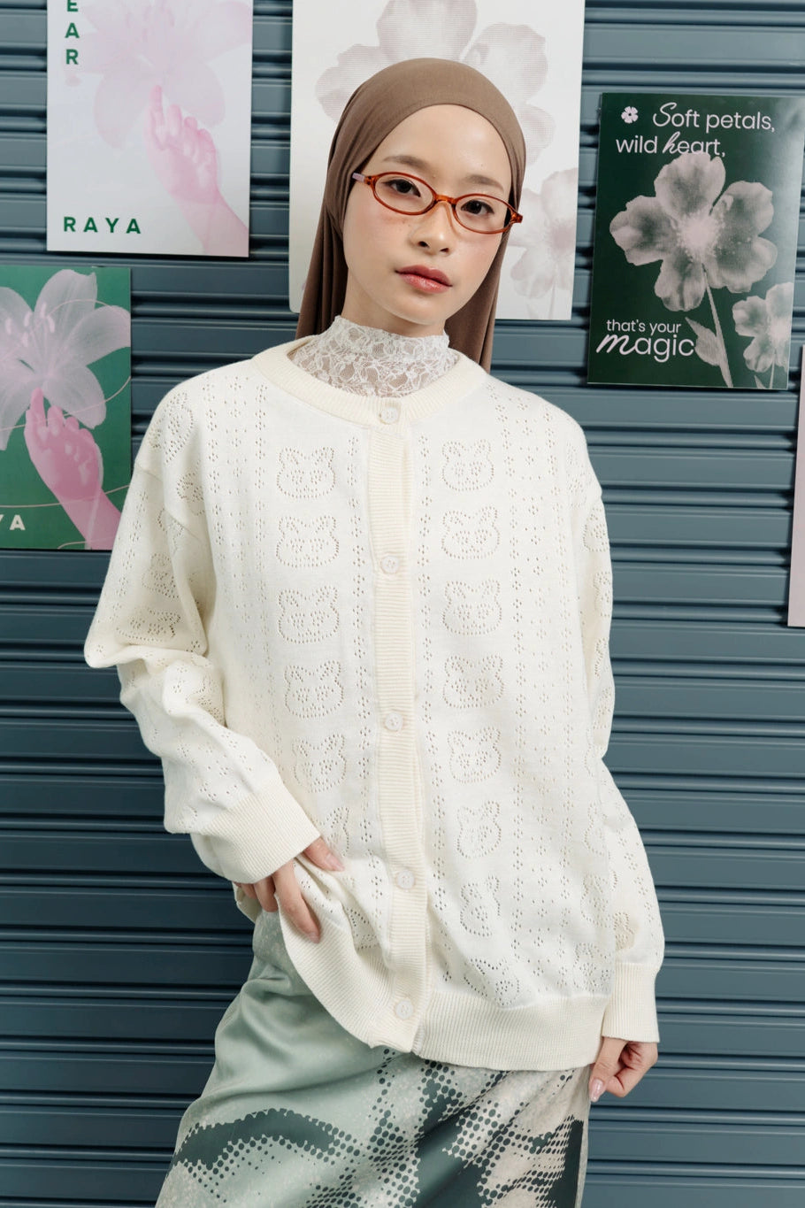Person wearing a white patterned cardigan by Tome Ame in front of a gray wall with floral posters.