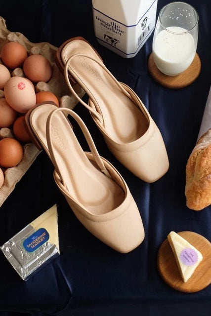 Pair of nude-colored flatshoes by Tangerine Marche on a table with eggs, milk, bread, and other items.