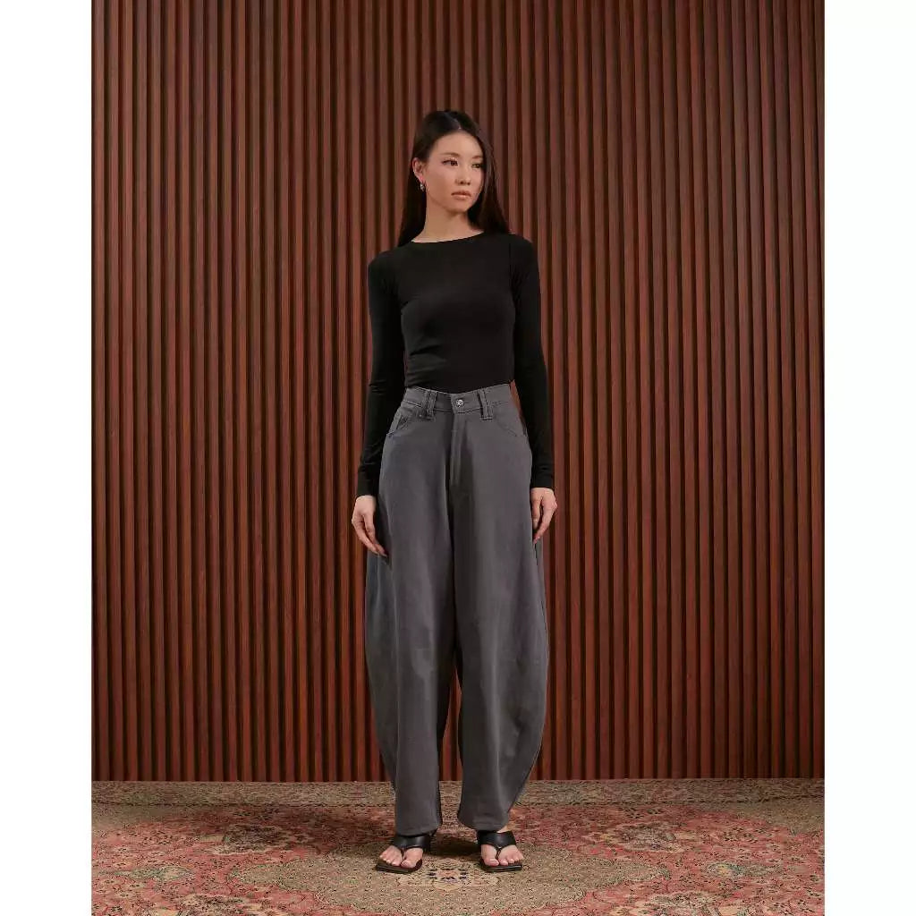 Woman wearing a black top and gray denim barrel pants standing against a brown striped wall.