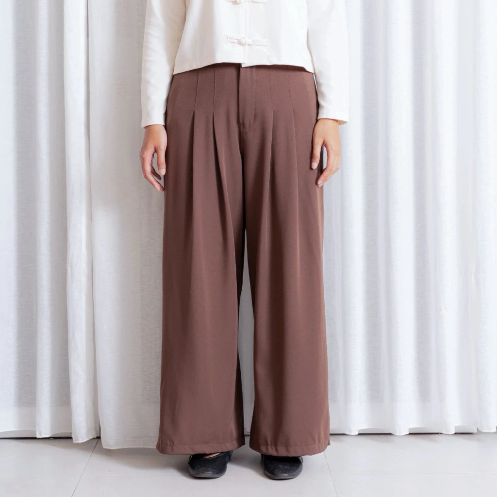 Person wearing brown wide-leg pants by notyourcompany and a white top against a white curtain background