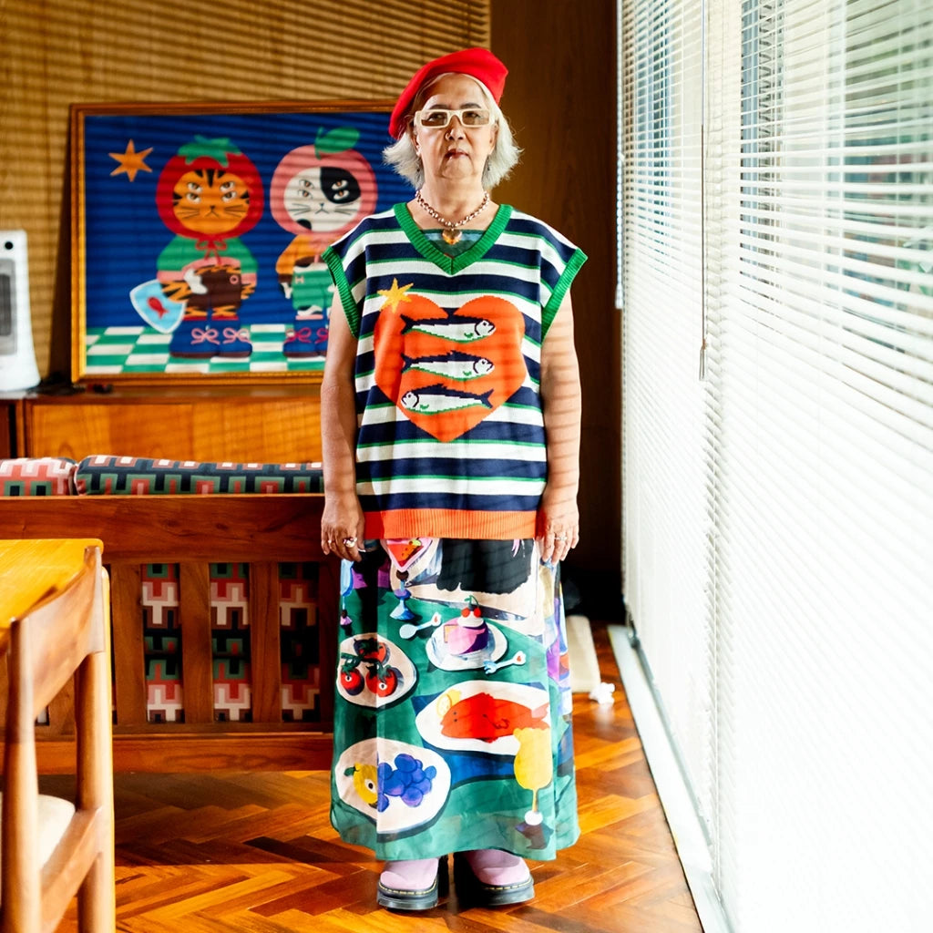 A person standing wearing "Dinner O Clock" knitted vest, a colorful knitted vest with a striped pattern and various food-themed graphics on it from Oh.Irv.