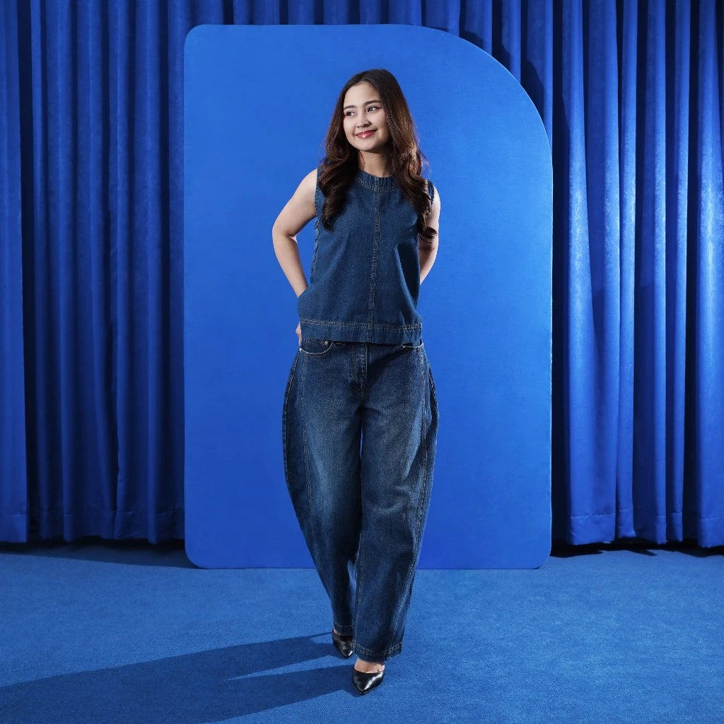Woman in a denim top and barrel pants standing against a blue curtain backdrop