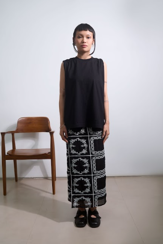 Person wearing a black sleeveless top by Tuff Puff and patterned black and white skirt standing against a white wall with a wooden chair.