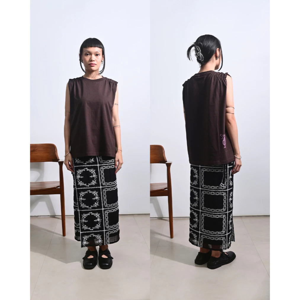 Person wearing a brown sleeveless top by Tuff Puff and black patterned skirt, standing in front of a white wall with two wooden chairs.