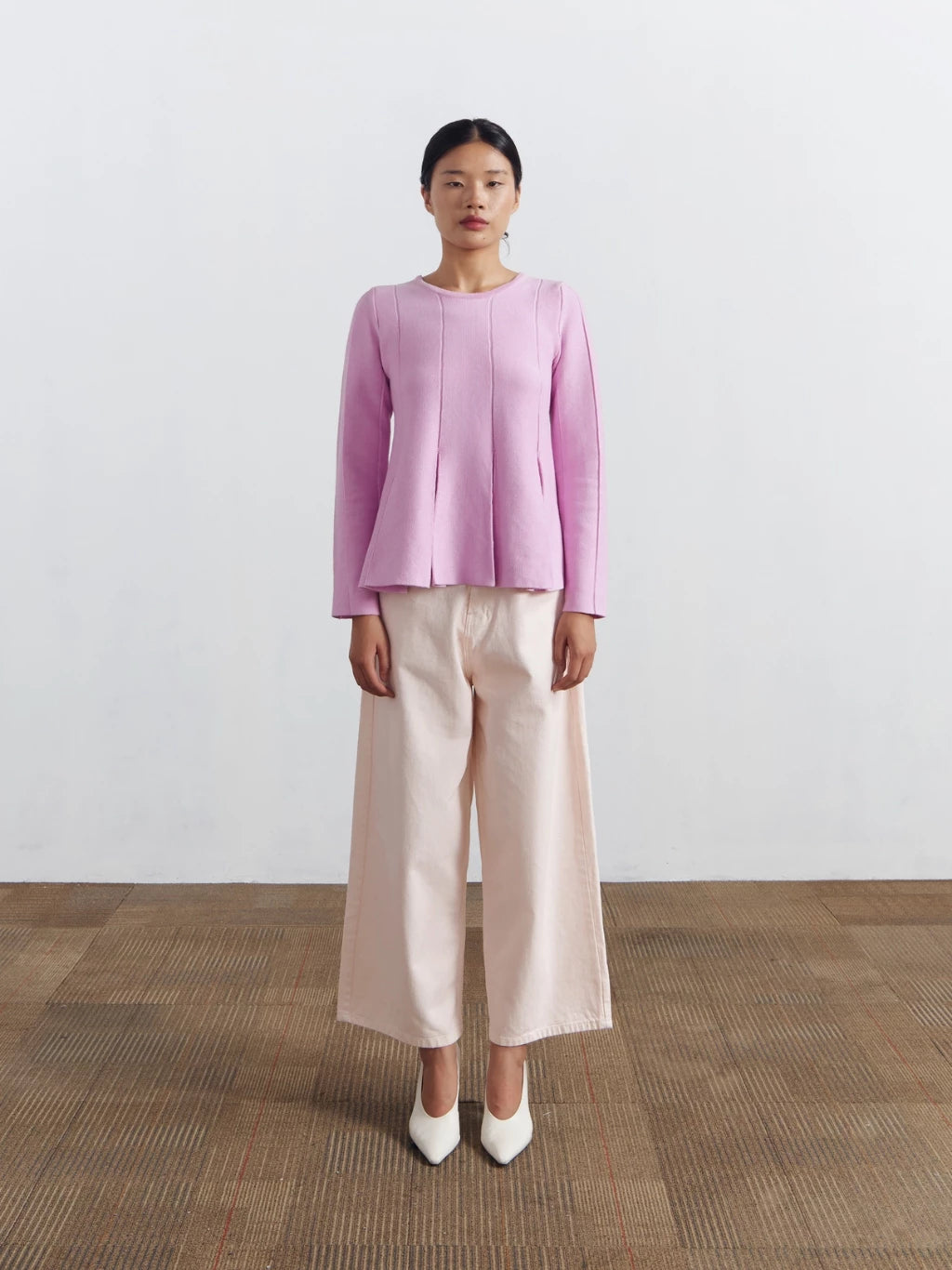 Woman wearing a pink longsleeved blouse by Achete de Nous and beige pants standing against a white wall.