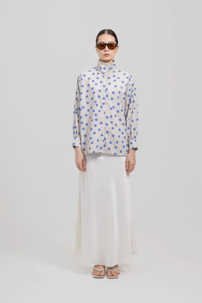 A model stands against a light gray studio background wearing a white long-sleeve mock-neck button-up shirt with a blue polka dots pattern by Tuff Puff and a long white skirt.