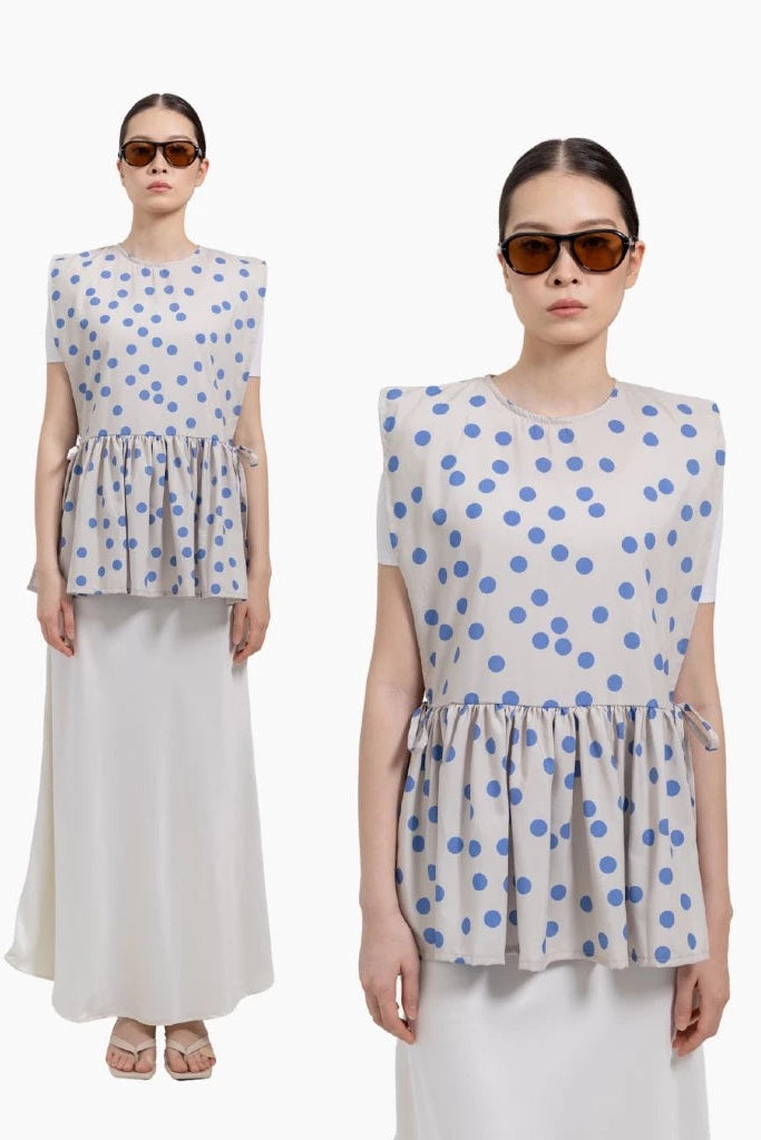A model is shown in two views wearing a sleeveless peplum top with blue polka dots by Tuff Puff on a light beige background, paired with a long white skirt. She is wearing dark sunglasses against a plain white studio background.