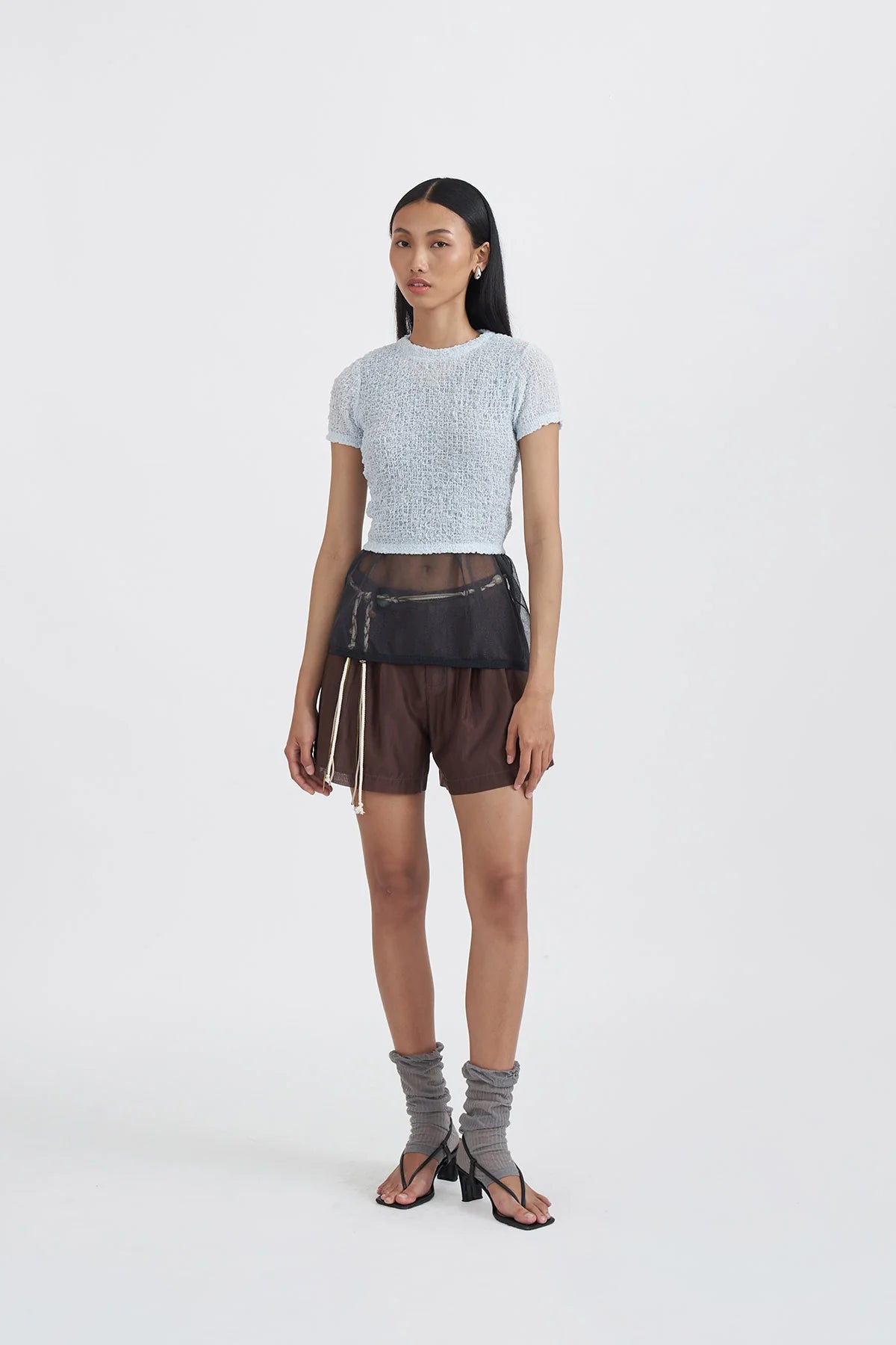 Woman wearing a light blue knit top by Callie Cotton and brown shorts with a sheer overlay on a white background