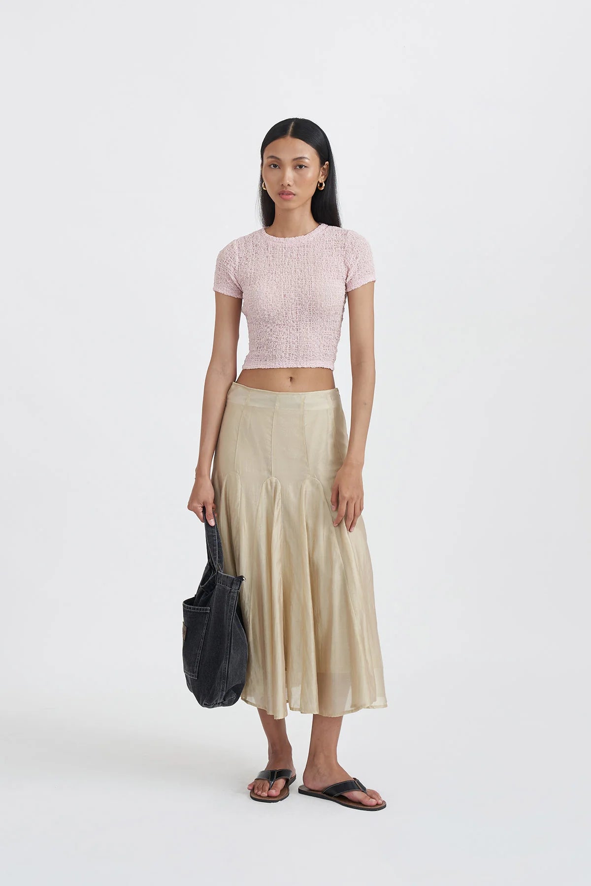 Woman wearing a pink knitted top by Callie Cotton and beige skirt with a black bag on a white background