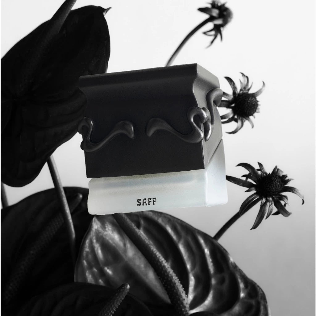 Black and white Saff perfume bottle from Saff & Co surrounded by dark leaves and flowers on a light background