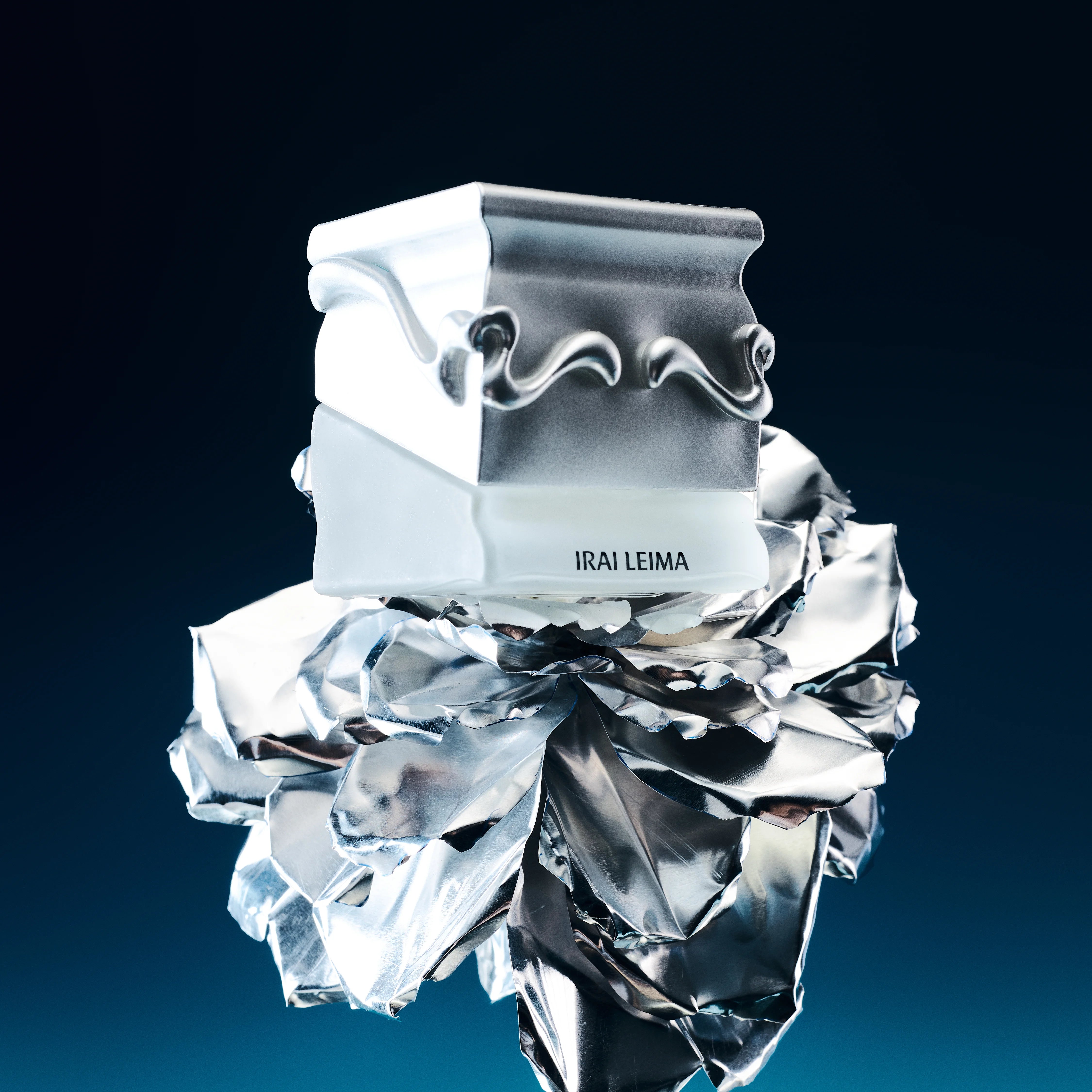 A glass Saff & Co. perfume bottle with a metallic design, the name 'IRAI LEIMA' inscribed on it, set against a blue background with a reflective effect below the bottle.