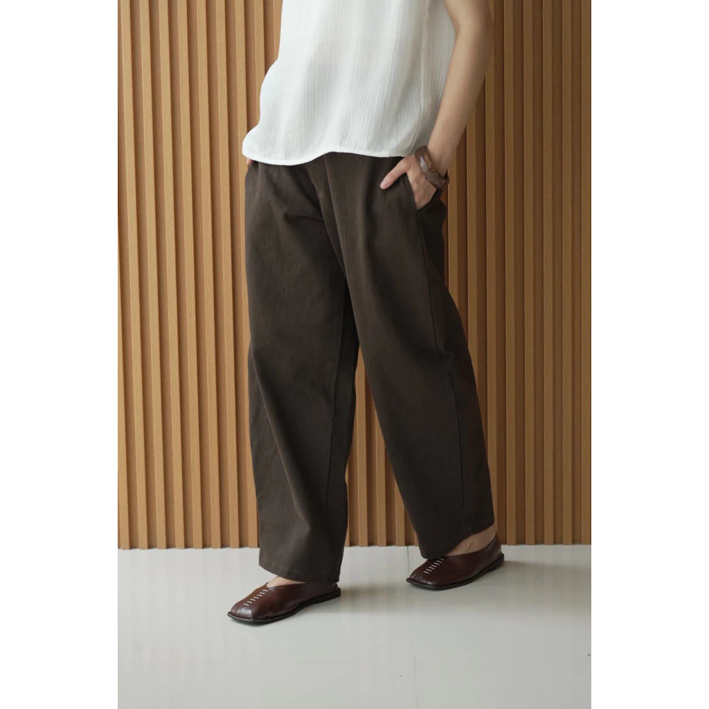 Person wearing a white top and dark brown barrel pants against a wooden paneled wall.