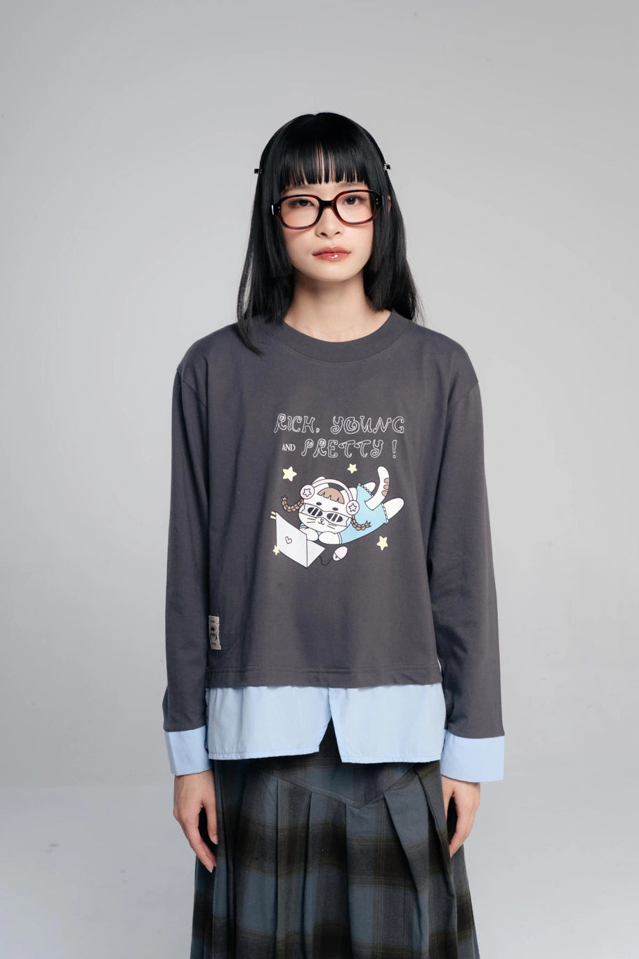 A model with black hair and glasses wearing a dark gray long-sleeve layered shirt by Tome Ame with a cartoon graphic and light blue hem detail, paired with a dark plaid skirt, against a plain gray background.