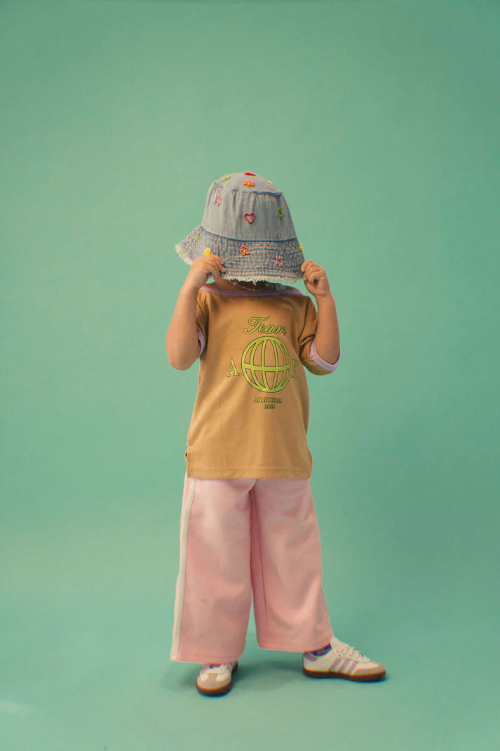 Child wearing a patterned bucket hat, yellow shirt with a globe design, and pink pants on a green background