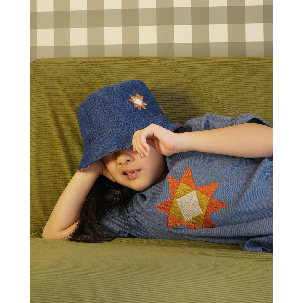 Kids wearing a blue denim hat and shirt with a sun design, lying on a green couch against a checkered wall.