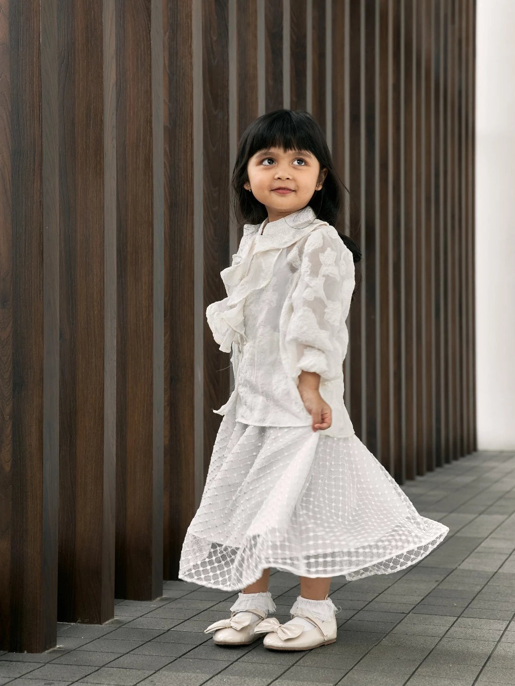 Young girl in a white top and white skirt standing against a wooden paneled wall.