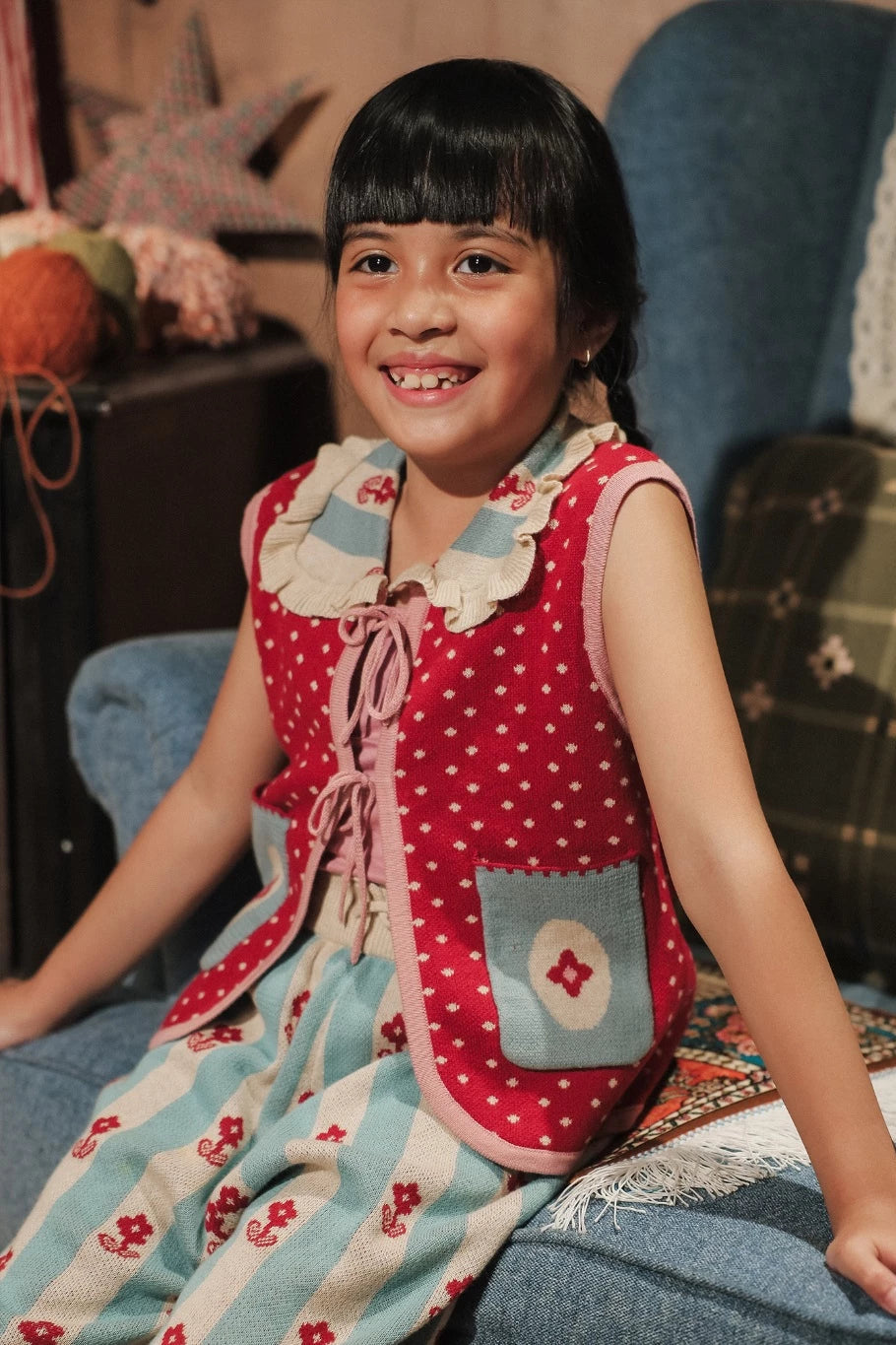 Young girl wearing a red vest with white polka dots and blue accents by Sabine & Heem kids clothing, sitting on a blue chair.