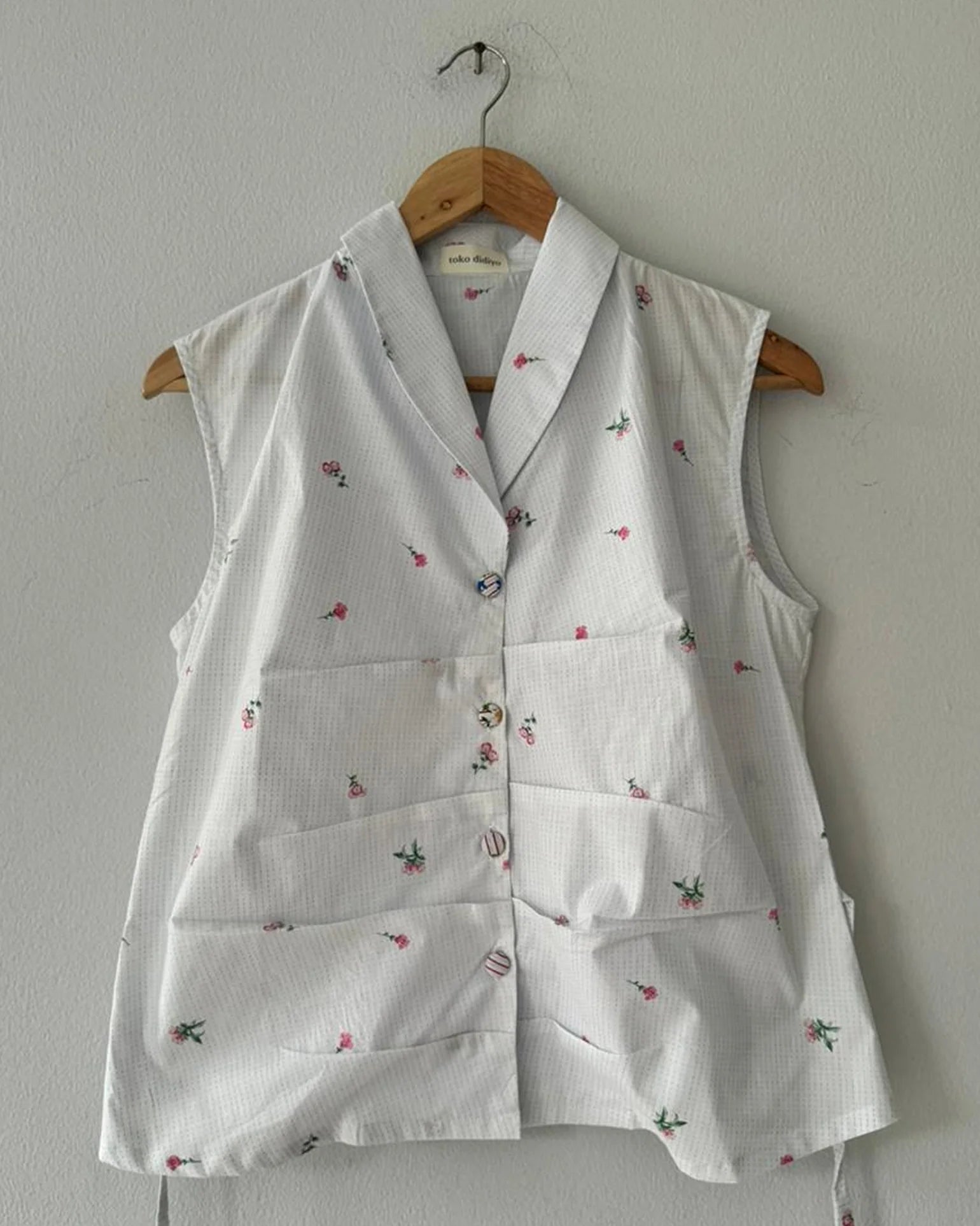 White sleeveless blouse by Toko Didiyo with floral patterns on a wooden hanger against a white wall.