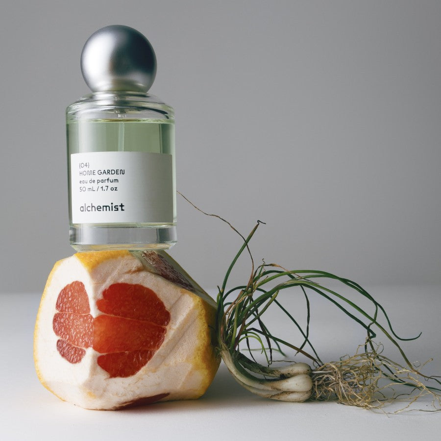 A bottle of 'Home Garden' Alchemist perfume with a silver cap, placed next to a slice of grapefruit and a sprig of plants on a white background.