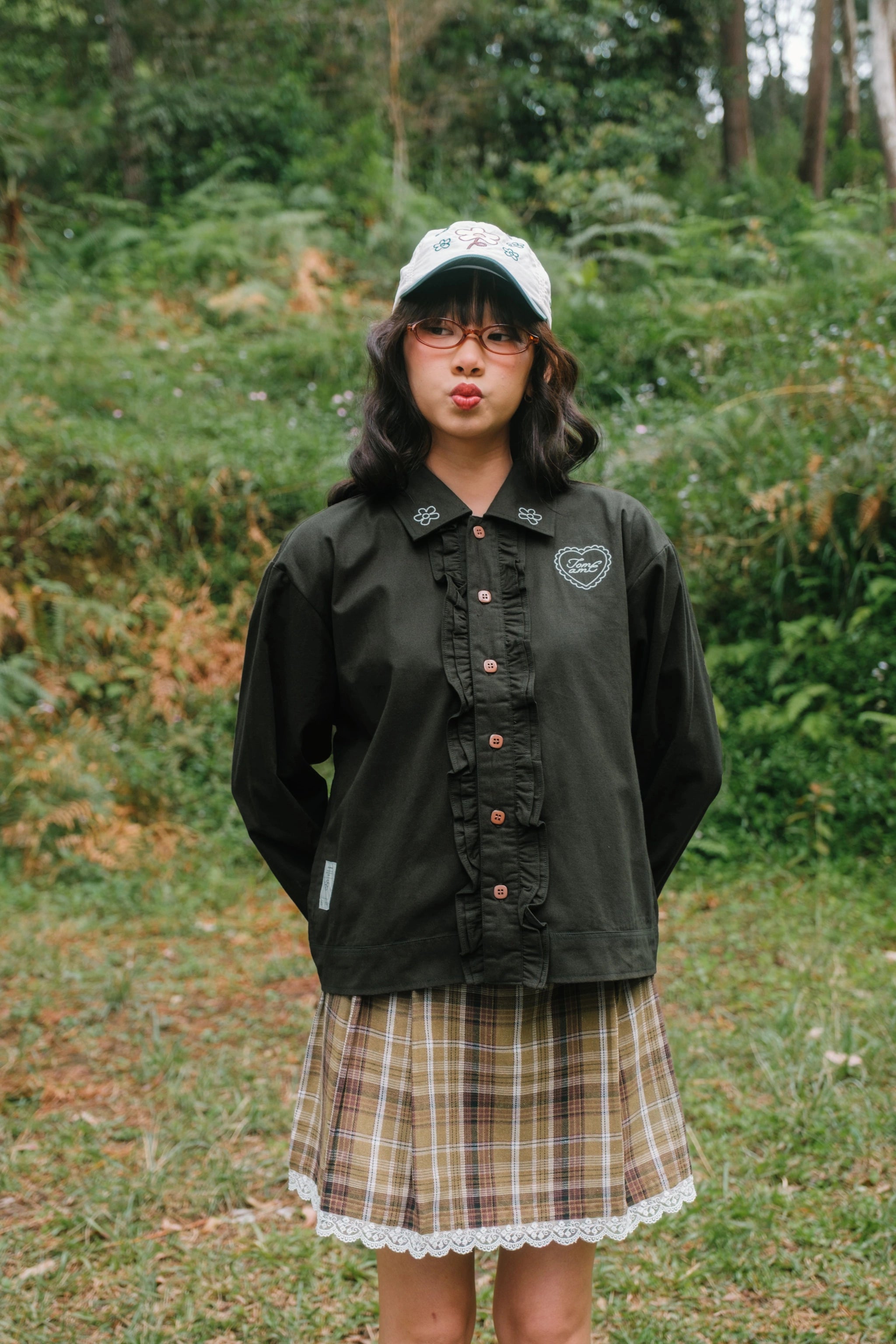 Person wearing a dark green jacket by Tome Ame and plaid skirt standing in a forest