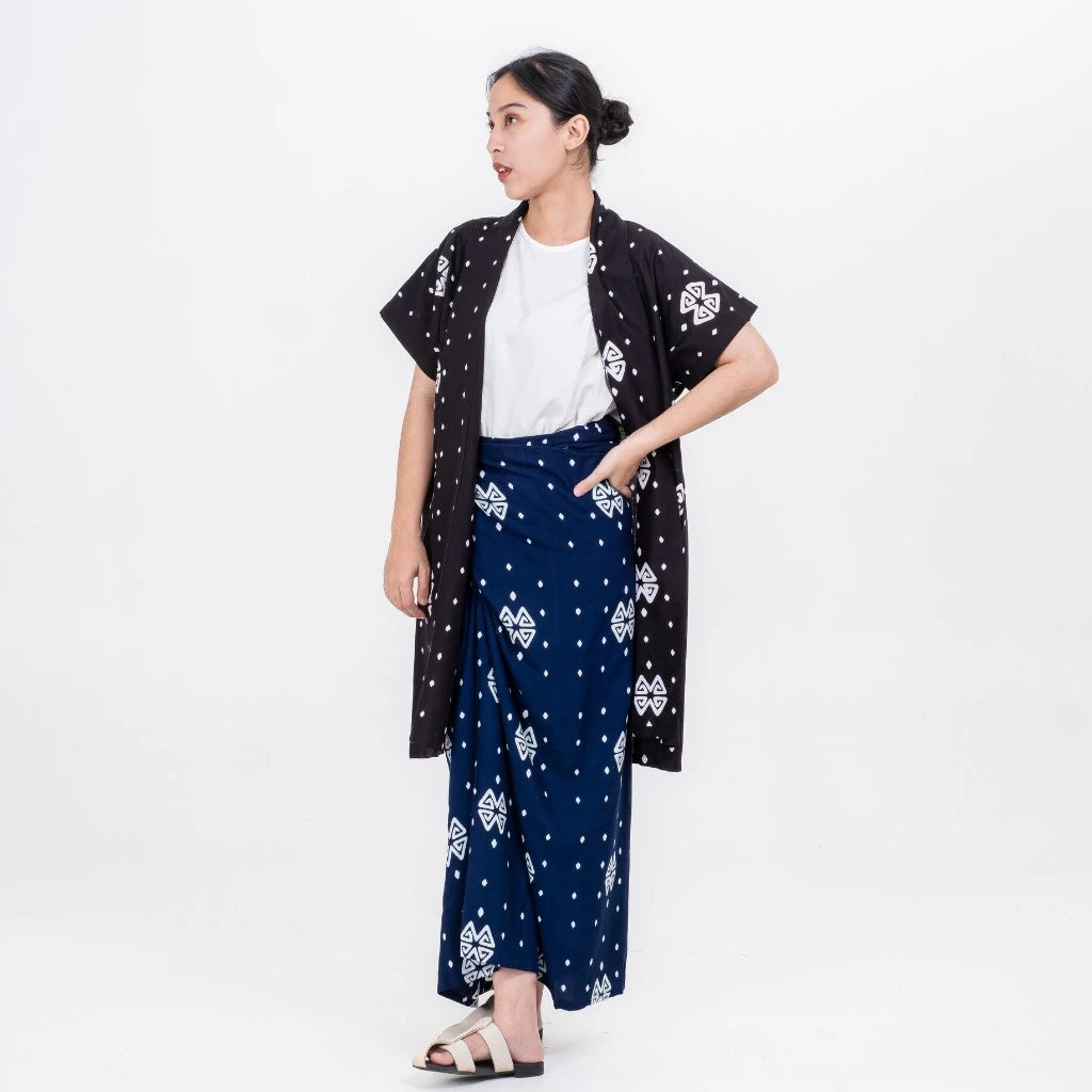 A woman standing wearing a traditional blue skirt with a white and blue floral pattern called Kaimana Skirt from Damakara, paired with a black traditional outer and a white top.