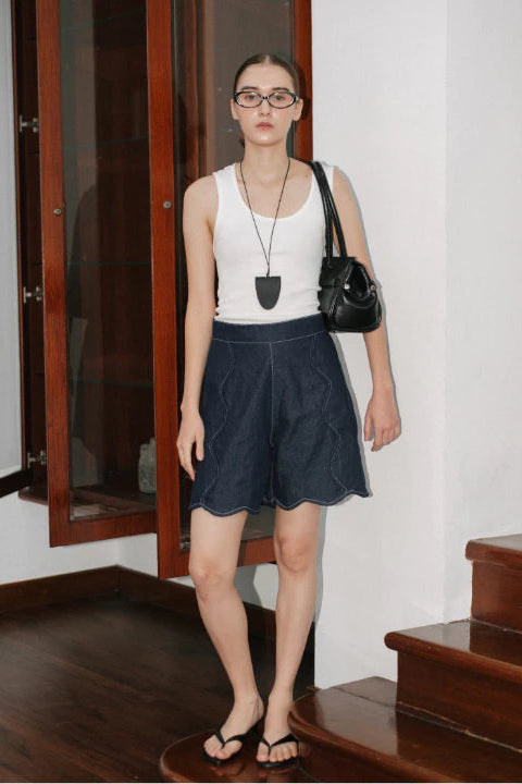 Woman wearing a white sleeveless top and dark blue jorts pants, standing indoors.