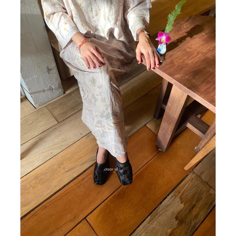 A woman wearing black embellished flatshoes with floral appliqué details, styled with a pastel floral-patterned dress, seated on wooden flooring while holding a small pink flower.