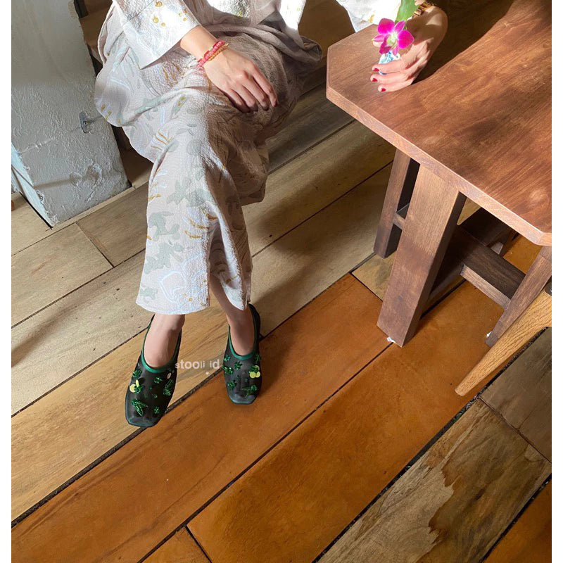 A woman wearing green embellished flatshoes with floral appliqué details, styled with a pastel floral-patterned dress, seated on wooden flooring while holding a small pink flower.
