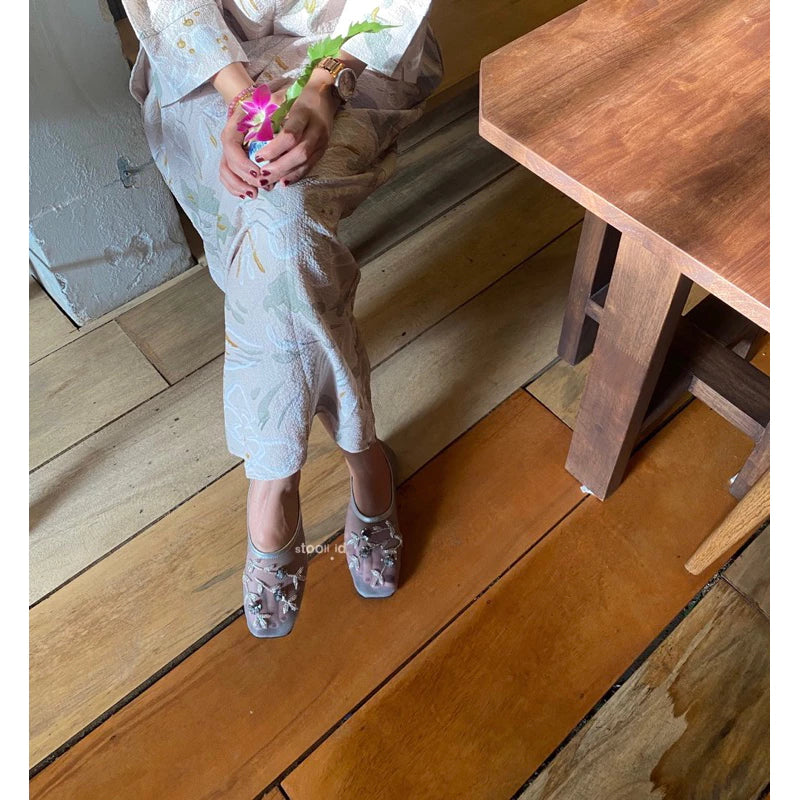 A woman wearing grey embellished flatshoes with floral appliqué details, styled with a pastel floral-patterned dress, seated on wooden flooring while holding a small pink flower.